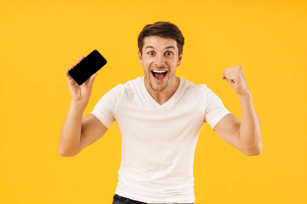 Feliz emocionado joven sorprendido en casual camiseta blanca utilizando el teléfono móvil aislado sobre fondo amarillo
. - Foto, imagen