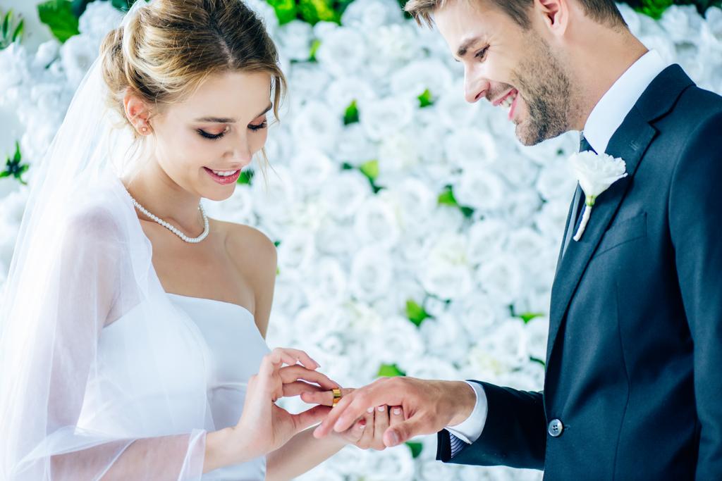 atractiva y sonriente novia poniendo anillo de bodas en el dedo - Foto, imagen