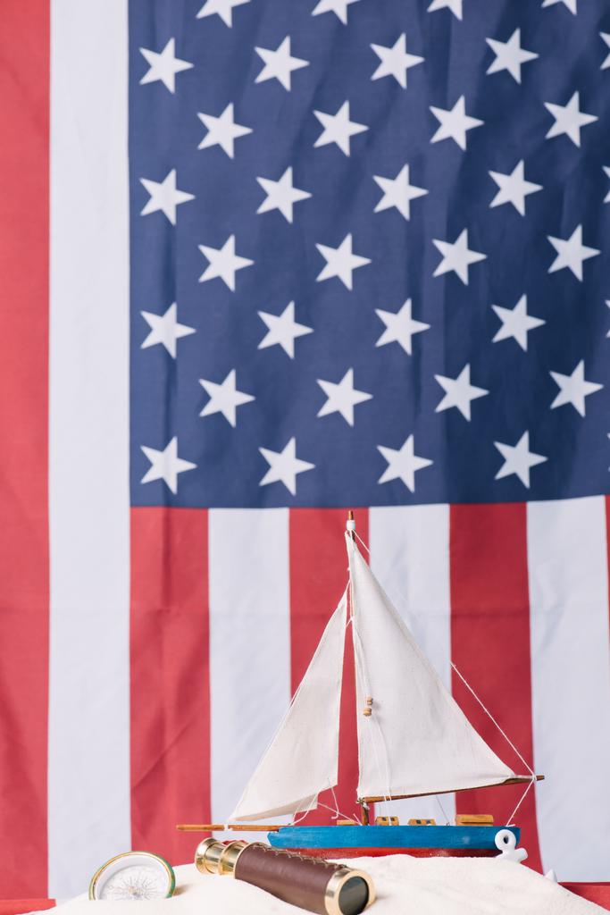 διακοσμητικό πλοίο σε λευκή άμμο κοντά στην πυξίδα και το τηλεσκόπιο στο φόντο της αμερικανικής εθνικής σημαίας - Φωτογραφία, εικόνα