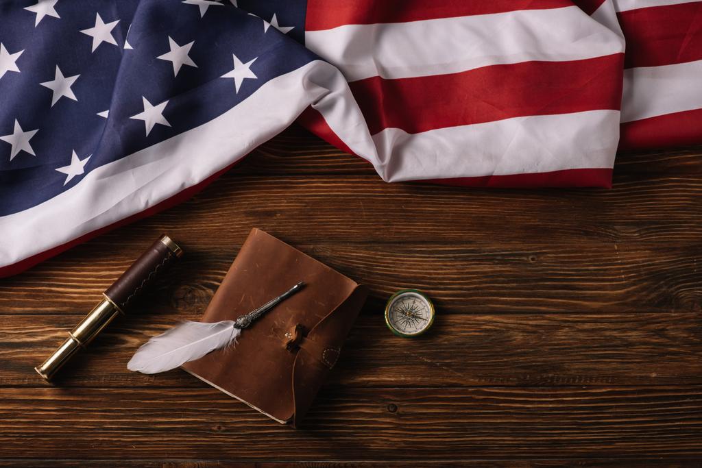 Draufsicht auf Lederheft, Teleskop, Feder und Kompass auf Holzoberfläche mit amerikanischer Nationalflagge - Foto, Bild