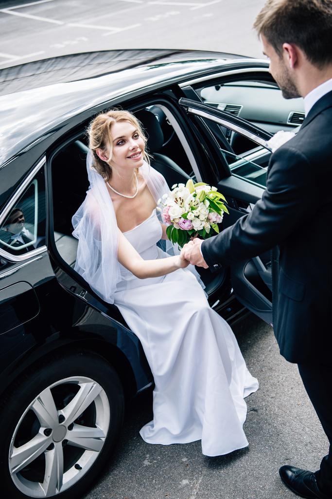 γαμπρός σε κοστούμι δίνοντας το χέρι σε ελκυστική και χαμογελαστή νύφη με ανθοδέσμη - Φωτογραφία, εικόνα