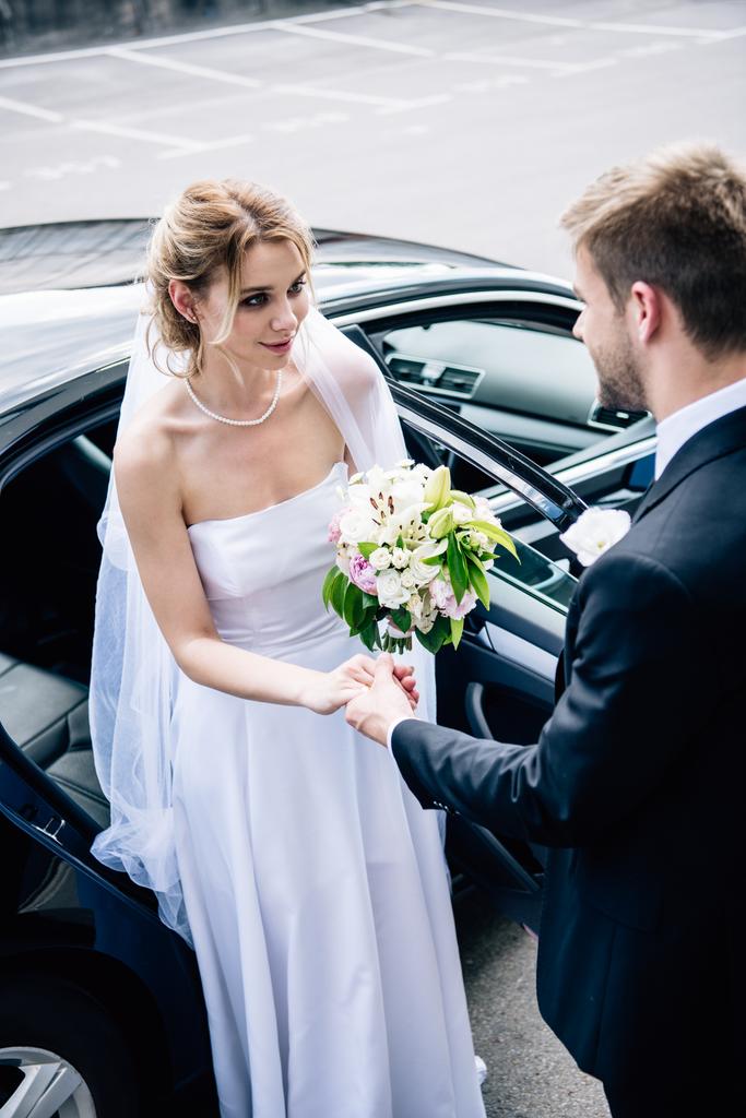 marié en costume donnant la main à la mariée attrayante et souriante avec bouquet
 - Photo, image