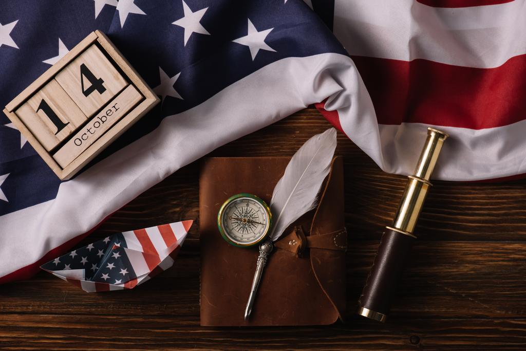 top view of wooden calendar with October 14 date, paper boat, nib, compass, telescope and leather notebook on wooden surface with American national flag - Photo, Image