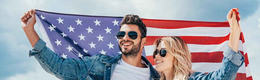 Panoramaaufnahme einer attraktiven Frau und eines gutaussehenden Mannes, die lächelnd die amerikanische Flagge halten  - Foto, Bild