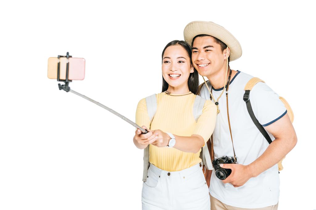 twee vrolijke Aziatische toeristen die selfie maken op smartphone met selfie stok geïsoleerd op wit - Foto, afbeelding