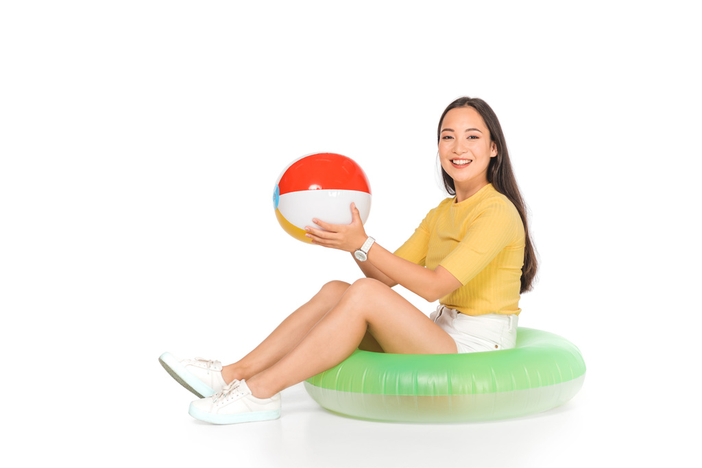 cheerful asian girl sitting on swim ring and holding beach ball on white background - Photo, Image