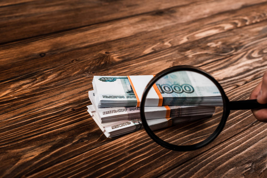 περικομμένη όψη του ανθρώπου κρατώντας μεγεθυντικό φακό κοντά στα Ρωσικά χρήματα στο τραπέζι  - Φωτογραφία, εικόνα