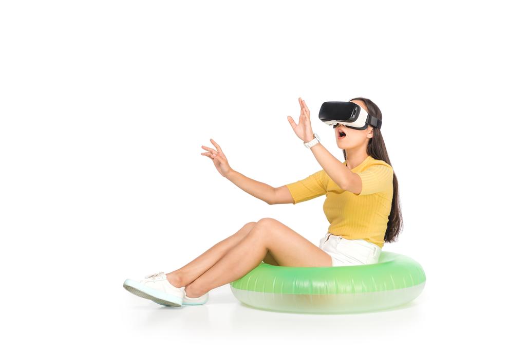 järkyttynyt aasialainen nainen käyttää virtuaalitodellisuus kuulokkeet ja eleitä istuessaan uimaan rengas valkoisella taustalla
 - Valokuva, kuva