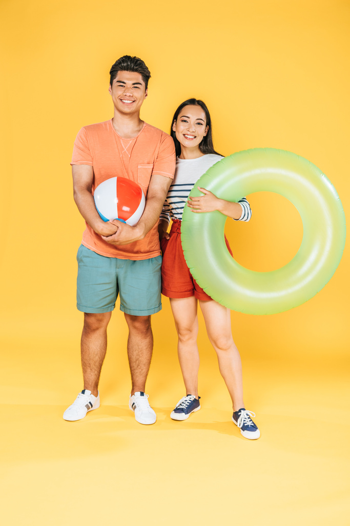 happy asian couple holding swim ring and beach ball while smiling at camera on yellow background - Photo, Image