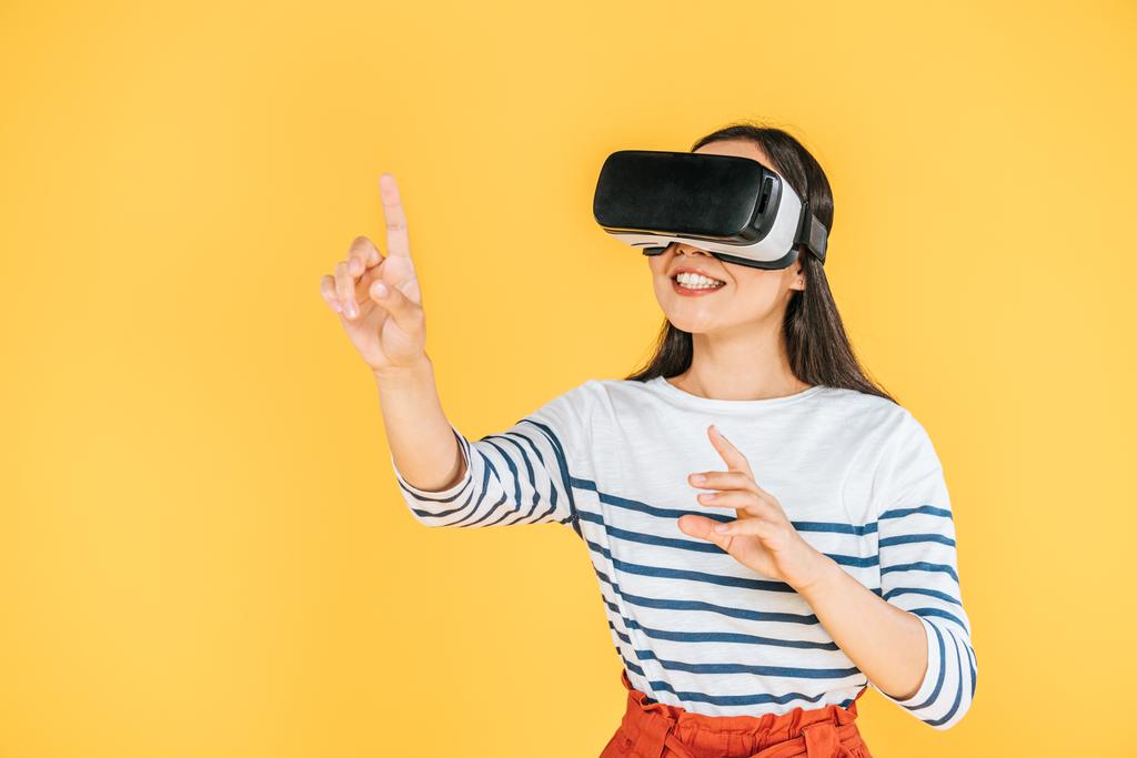 glimlachend meisje gebaren tijdens het gebruik van Virtual Reality headset geïsoleerd op geel - Foto, afbeelding