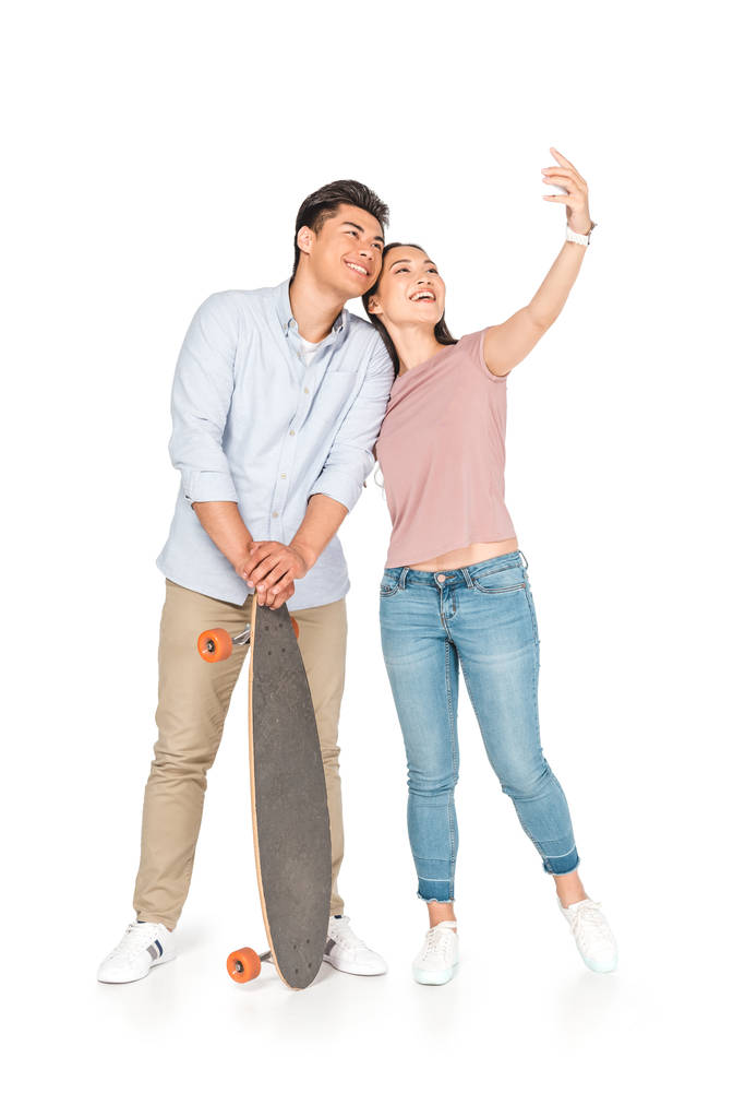 beyaz arka plan üzerinde longboard tutan yakışıklı erkek arkadaşı ile selfie çeken mutlu asyalı kız - Fotoğraf, Görsel