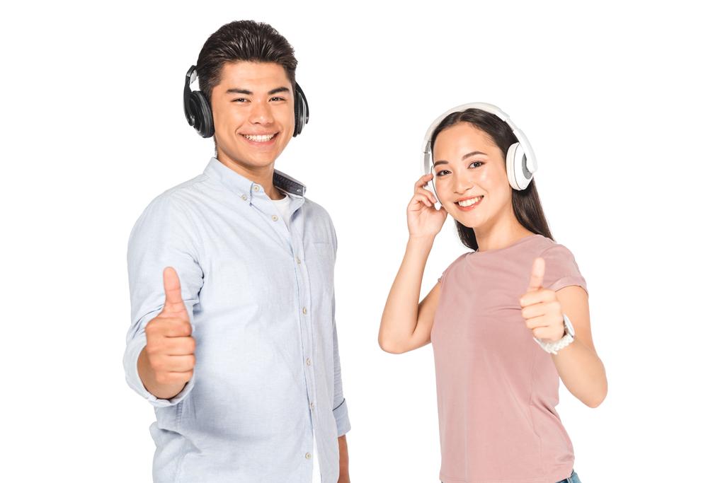 χαμογελαστός Ασιάτης και γυναίκα που δείχνει τους αντίχειρες πάνω ακούγοντας μουσική στα ακουστικά και κοιτώντας την κάμερα απομονωμένη σε λευκό - Φωτογραφία, εικόνα