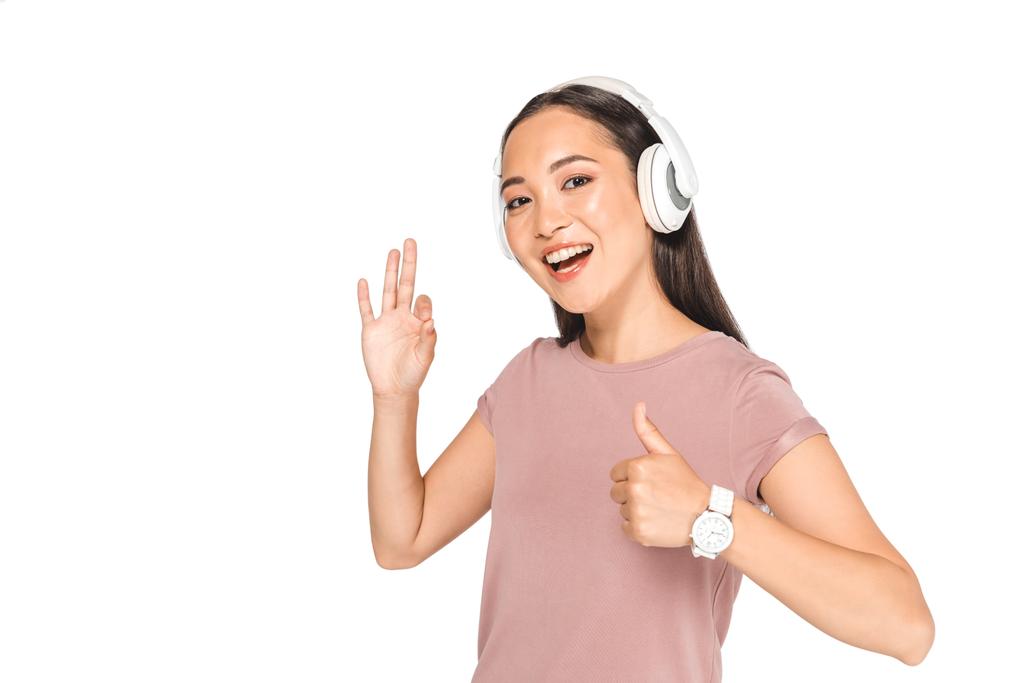bonita mujer asiática escuchando música en los auriculares, mostrando el pulgar hacia arriba y el gesto bien mientras sonríe a la cámara aislada en blanco
 - Foto, Imagen
