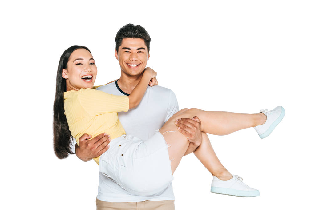 vrolijke Aziatische man houdt gelukkige vriendin op handen en glimlachend op de camera geïsoleerd op wit - Foto, afbeelding