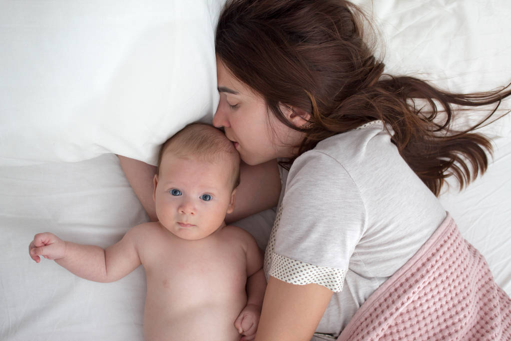 Piękna brunetka dziewczyna całuje jej dziecko. Są w sypialni - Zdjęcie, obraz