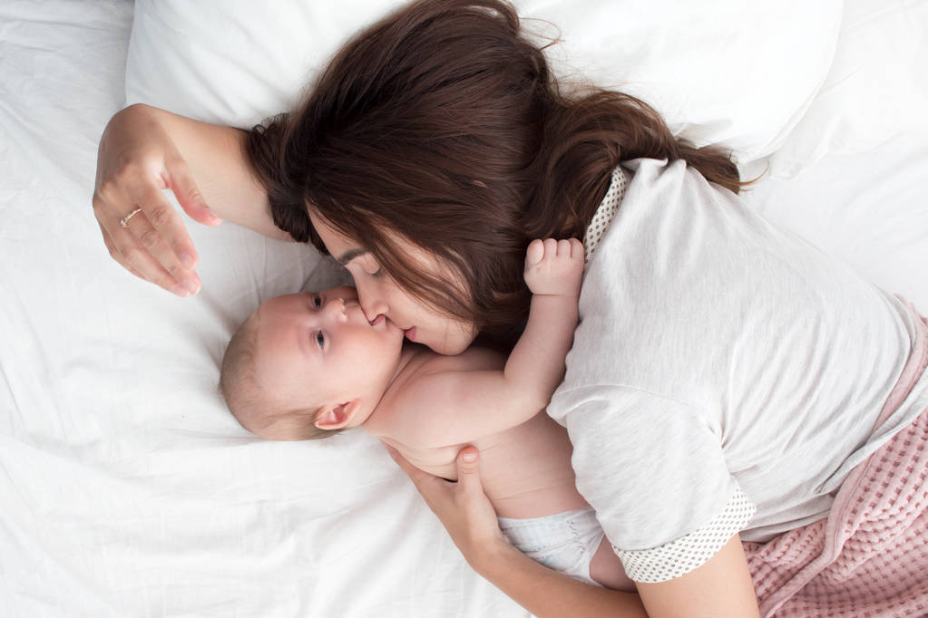 Όμορφη μελαχρινή κορίτσι που φιλάει το μωρό της. Είναι στην κρεβατοκάμαρα. - Φωτογραφία, εικόνα