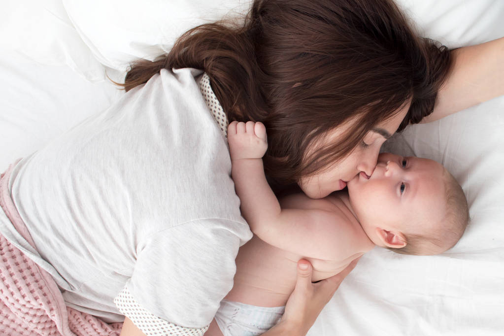 Красива дівчина брюнетка цілує свою дитину. Вони в спальні
 - Фото, зображення