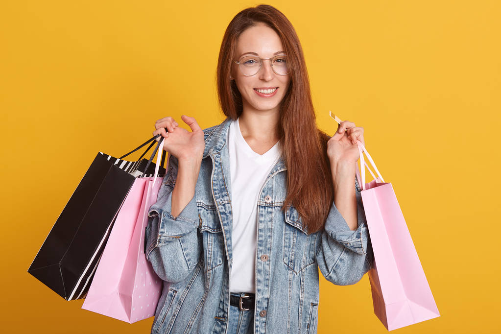 Foto einer charmanten Frau, die Einkaufstüten in den Händen hält und direkt in die Kamera blickt, auf gelbem Hintergrund. schöne junge kaukasische Shopperin lächelt glücklich, Dame hat dunkle glatte Haare. - Foto, Bild