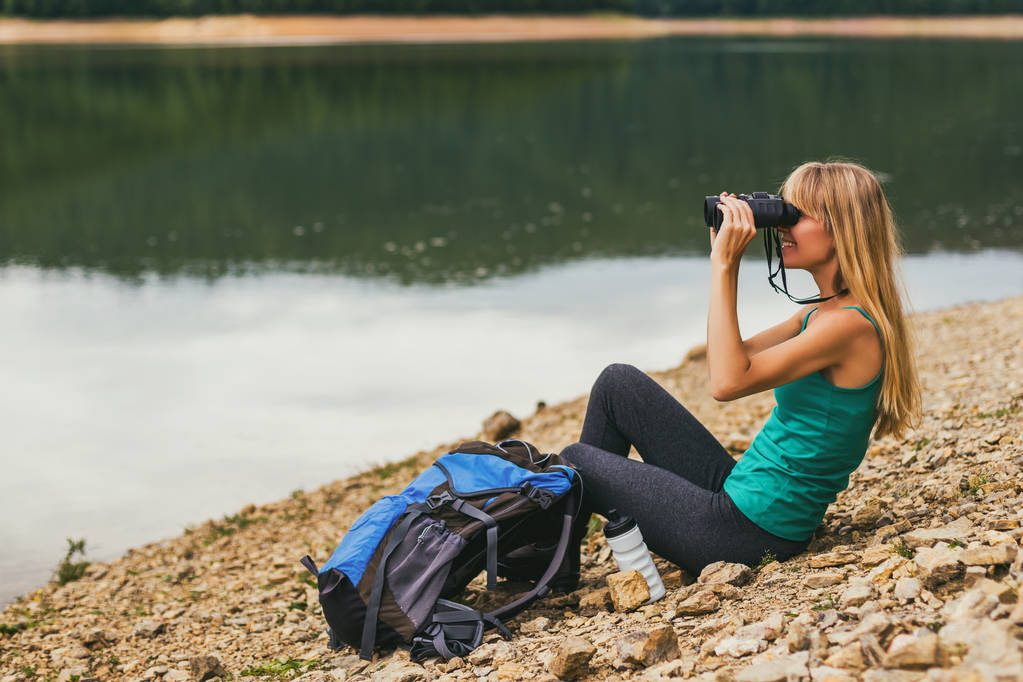 Femme randonneur utilisant des jumelles tout en étant assis au bord du lac
. - Photo, image