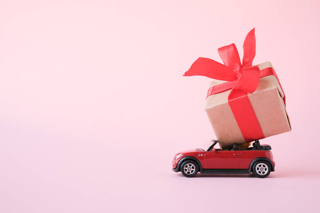 coche de juguete rojo entrega caja de regalo con cinta roja sobre fondo rosa. Lugar para el texto. Ventas, cumpleaños, Navidad. San Petersburgo Rusia 16 julio 2019
. - Foto, imagen