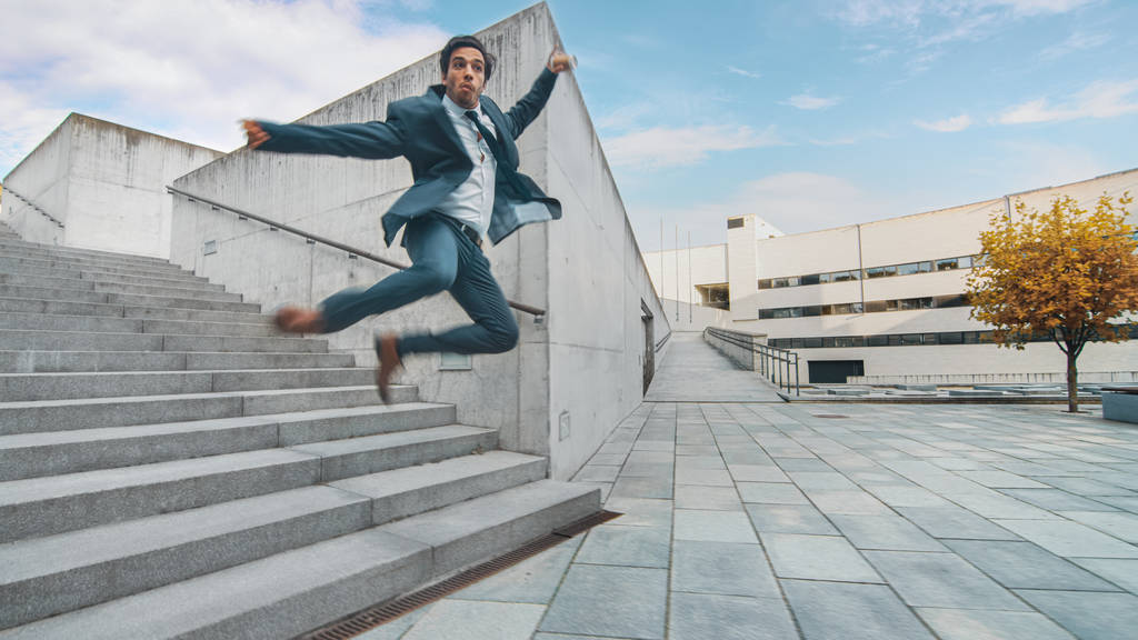 Веселый и счастливый бизнесмен в костюме держит кофе и прыгает во время прогулки по лестнице. Сцена снята в городском бетонном парке рядом с бизнес-центром. Солнечный
. - Фото, изображение
