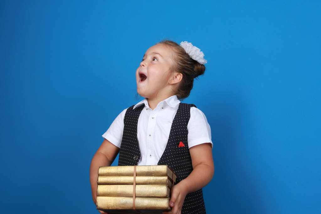 Surprised excited girl wearing school uniform, holding stack of golden books and looking at copy space with open mouth. Back to school, education, elementary school concept - Photo, Image
