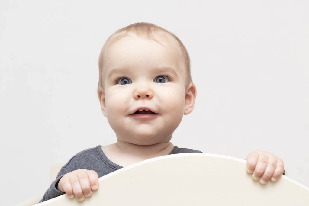 Porträt europäisch-kaukasisch glücklich lächelndes Kleinkind mit blauen Augen auf weißem Hintergrund, das über die Kamera blickt, die sich am Gitterbett festhält. Alter - 10 Monate - 1 Jahr - Foto, Bild