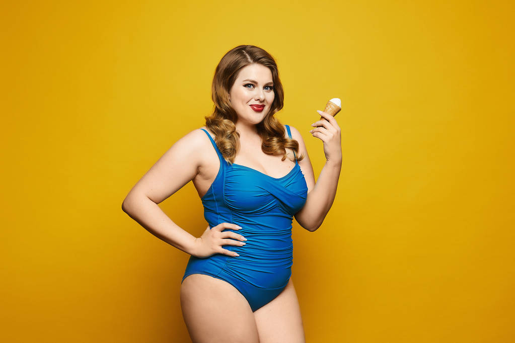 Beautiful and sexy plus size model girl, with stylish hairstyle and red lips, in fashionable blue swimsuit eating tasty ice cream and posing at the yellow background, isolated - Photo, Image
