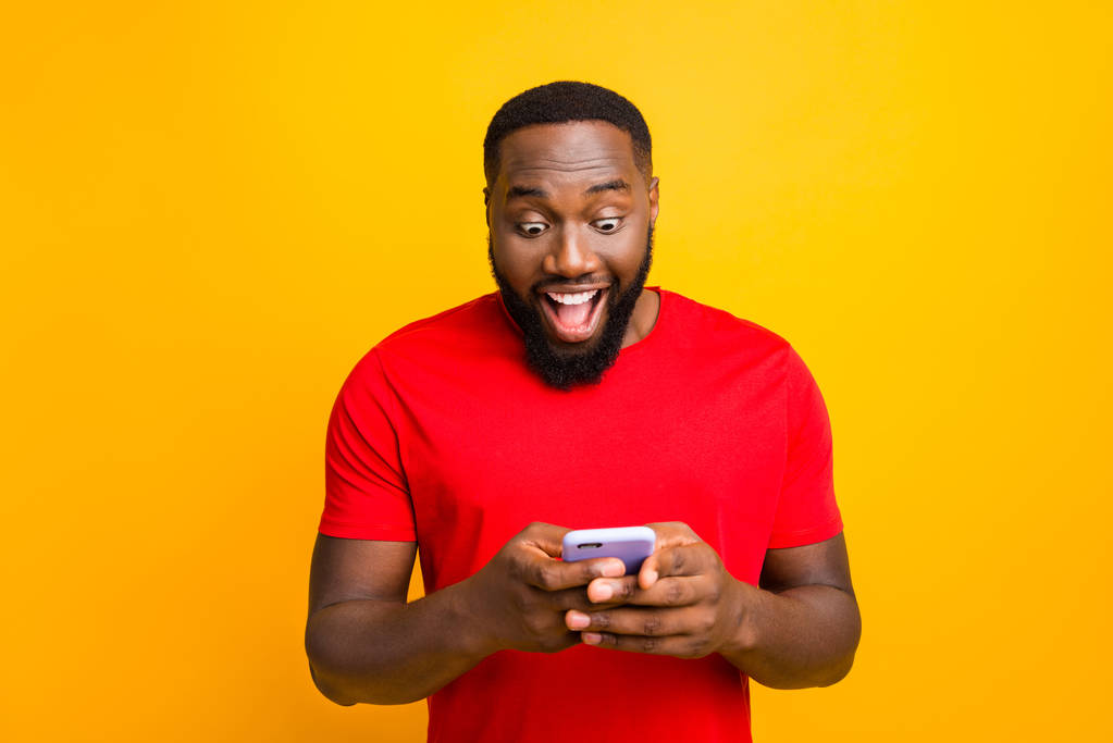 Photo of impressed black man watching something delightful on his phone wearing red t-shirt while isolated with yellow background - Photo, Image