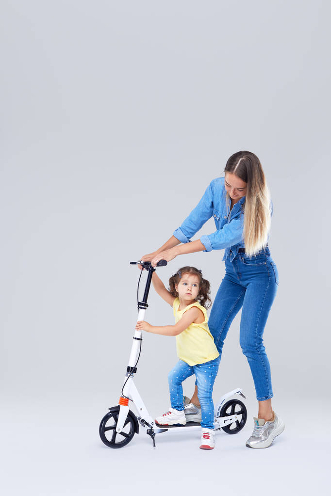 Mujer joven y niña de pie en scooter y monopatín. Mamá enseña a su hija a montar, divertirse y mirar alegremente en la cámara sobre fondo gris
 - Foto, imagen