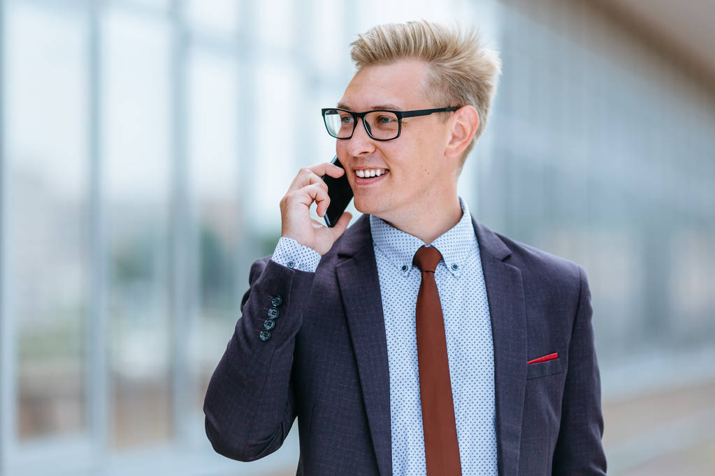 オフィスビルの前で携帯電話で話すメガネをかけた若いハンサムな白人ビジネスマン。屋外でスマートフォンを使用してトレンディな服を着た男性エグゼクティブマネージャー。現代技術の概念 - 写真・画像