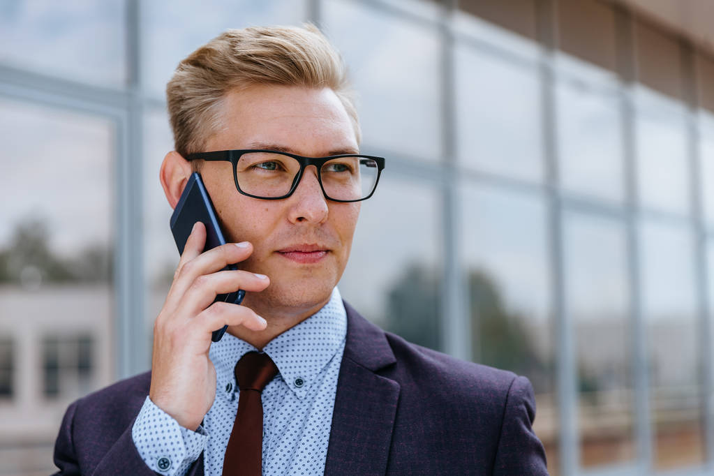 Młody przystojny kaukaski biznesmen w okularach rozmowy na telefon komórkowy przed budynkiem biurowym. Męski kierownik wykonawczy w modne ubrania za pomocą smartfona na zewnątrz. Nowoczesna koncepcja technologiczna - Zdjęcie, obraz