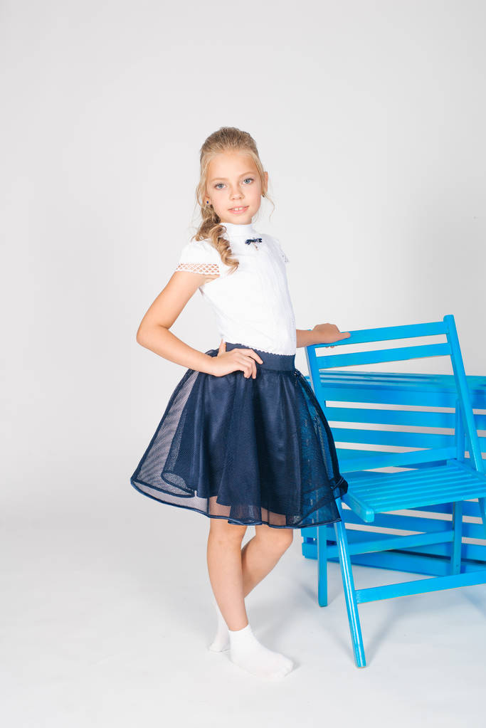 Cute Girl z długim blond kręcone włosy w szkole mody ubrania z niebieskim krzesłem na białym tle - Zdjęcie, obraz