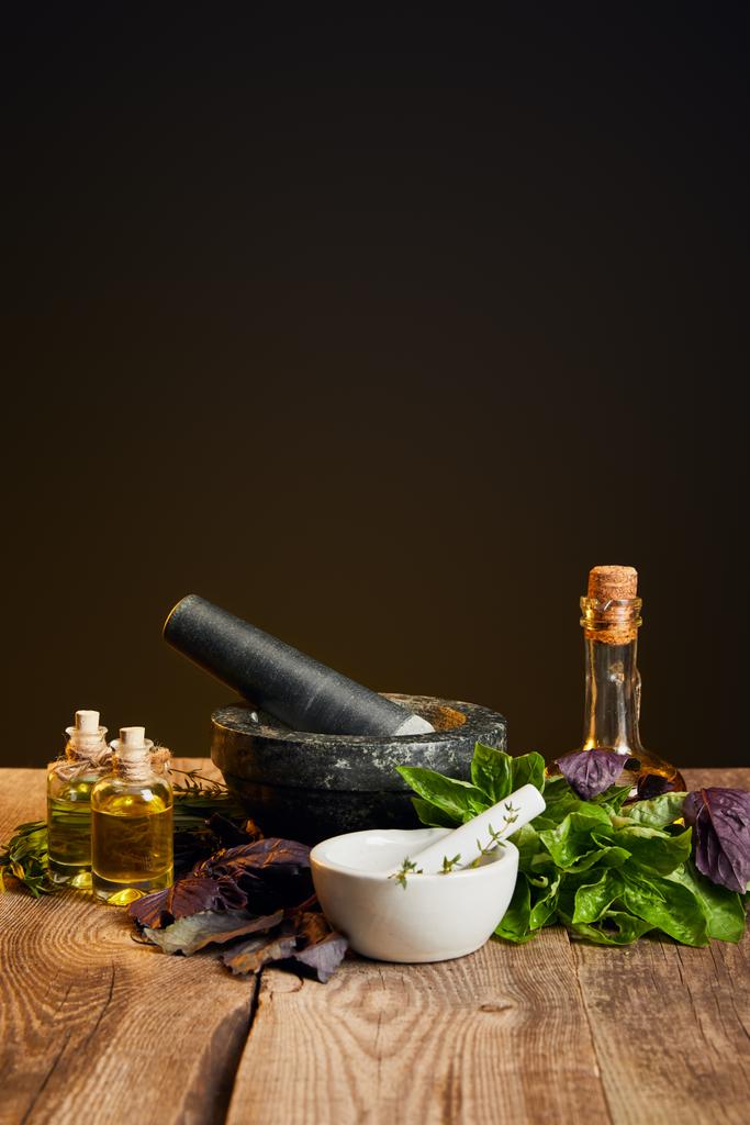 moździerze z tłuczek i butelek z olejem w pobliżu świeżych ziół na drewnianym stole izolowane na czarno - Zdjęcie, obraz