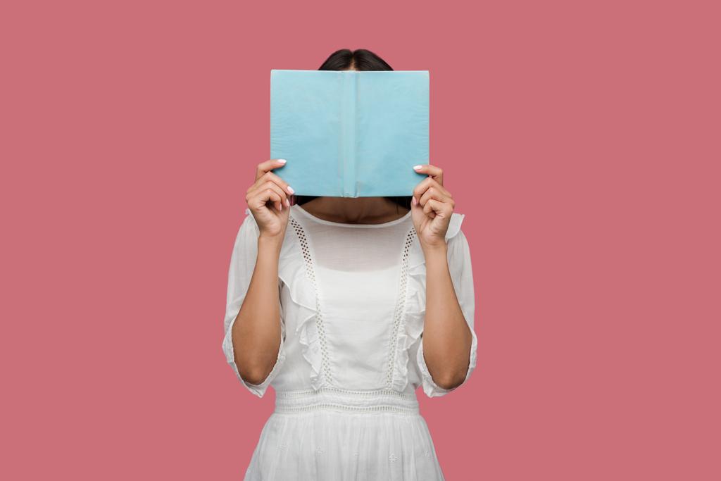 mujer joven en vestido cubriendo la cara con libro azul aislado en rosa
  - Foto, Imagen