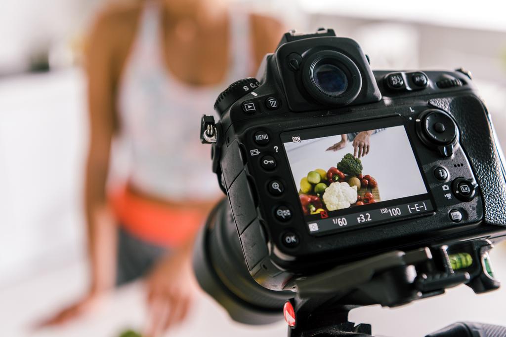 Selektiver Fokus der Digitalkamera mit Sportlerin, die in der Nähe von Gemüse auf dem Bildschirm gestikuliert  - Foto, Bild
