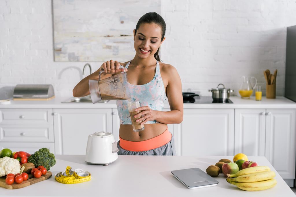 happy sportswoman pouring smoothie into glass near fruits and vegetables  - Photo, Image