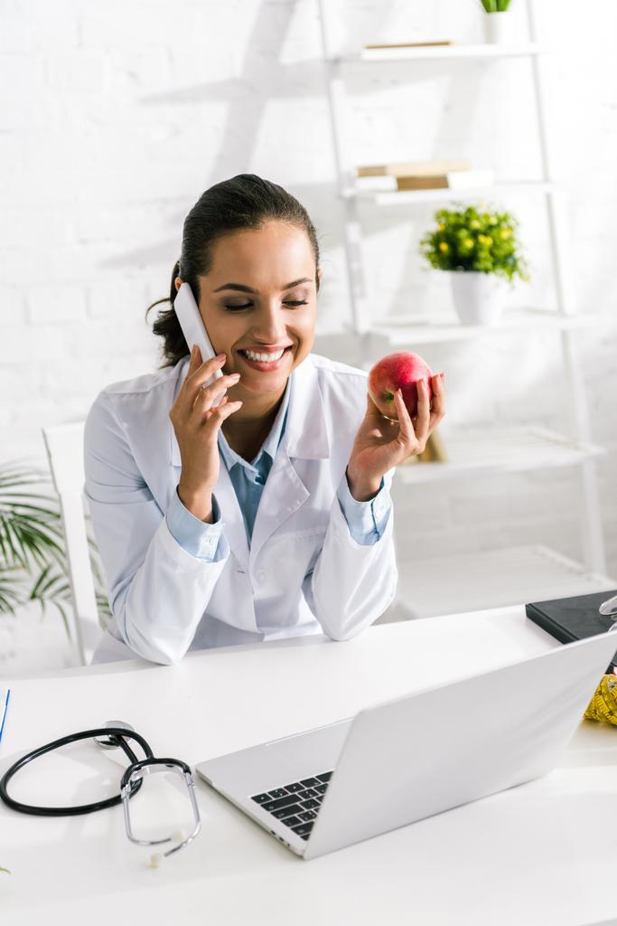 iloinen ravitsemusterapeutti puhuu älypuhelimella ja pitää omena klinikalla
  - Valokuva, kuva