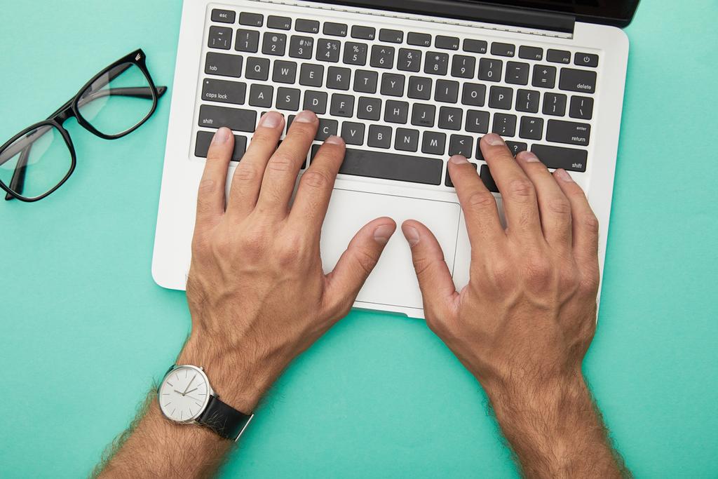 vista superior del hombre escribiendo en el ordenador portátil cerca de gafas aisladas en turquesa
 - Foto, imagen