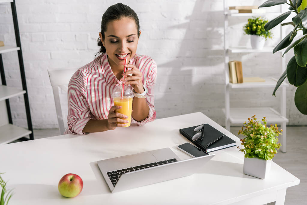 Szczęśliwa kobieta trzyma plastikową filiżankę z sokiem pomarańczowym w pobliżu laptopa i jabłko  - Zdjęcie, obraz