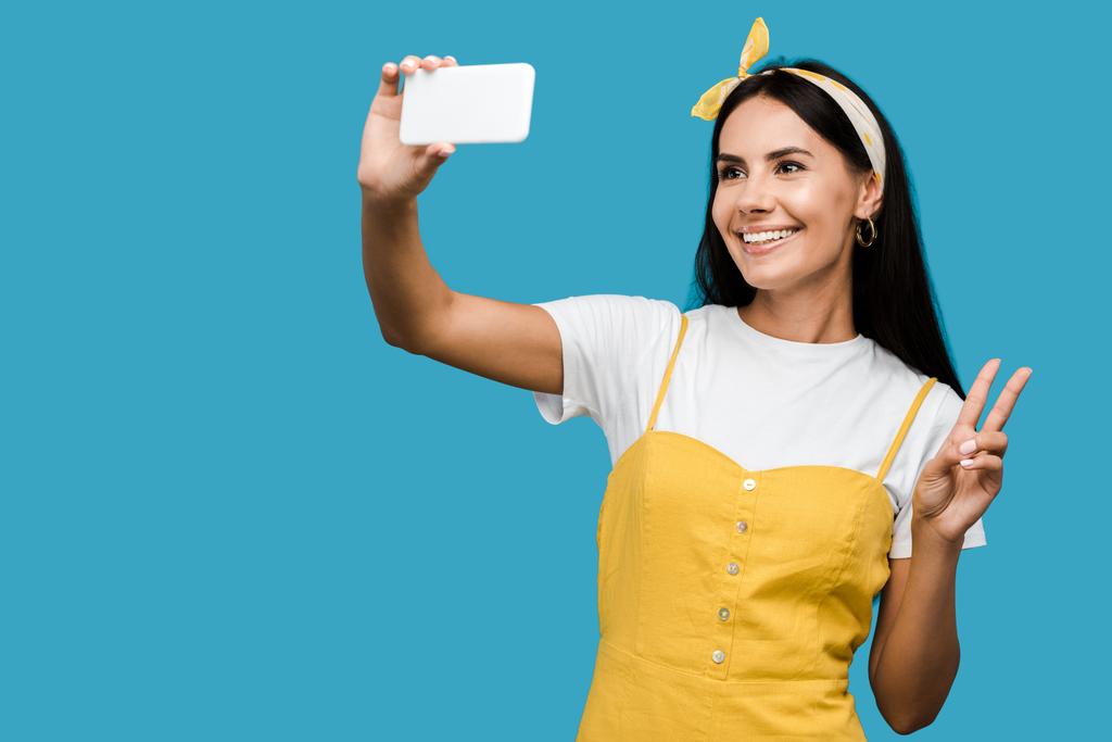 enfoque selectivo de la mujer alegre tomando selfie en el teléfono inteligente, mientras que muestra signo de paz aislado en azul
   - Foto, imagen
