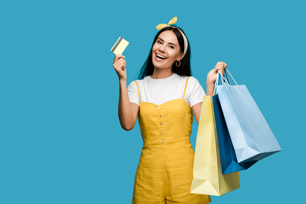 šťastná mladá žena držící kreditní kartu a nákupní tašky izolované na modrém  - Fotografie, Obrázek