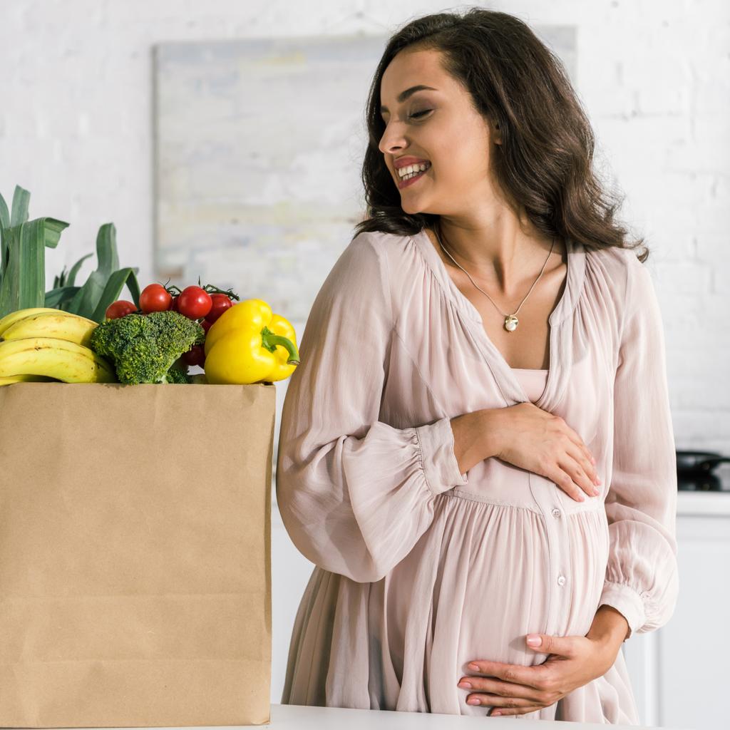 vrolijke zwangere vrouw op zoek naar papieren tas met boodschappen terwijl het aanraken van de buik  - Foto, afbeelding