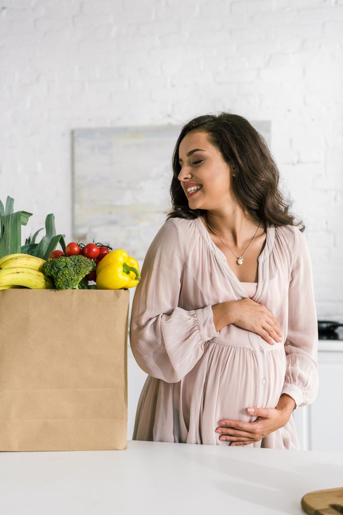 glückliche schwangere Frau blickt auf Papiertüte mit Lebensmitteln, während sie Bauch berührt  - Foto, Bild