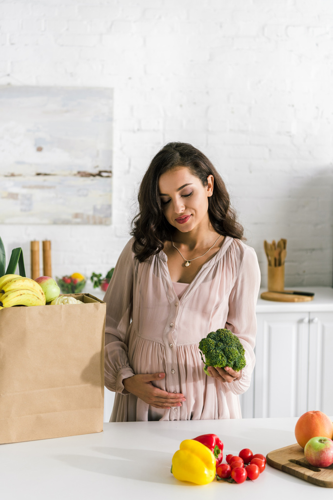 gelukkige zwangere vrouw houdt broccoli in de buurt van groenten in papieren zak  - Foto, afbeelding