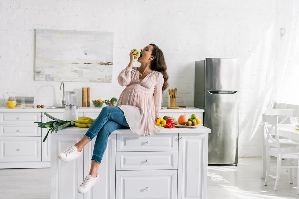 glückliche schwangere Frau isst Apfel neben leckerem Essen auf dem Tisch  - Foto, Bild