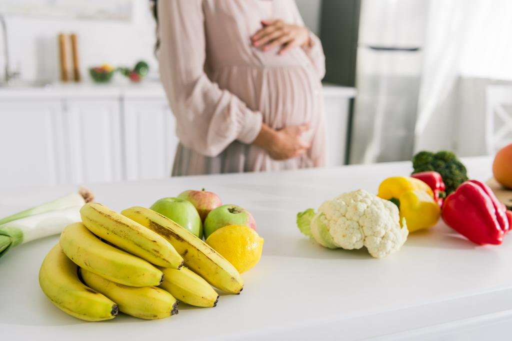 テーブルの上の果物の近くに立っている妊婦のトリミングされたビュー  - 写真・画像