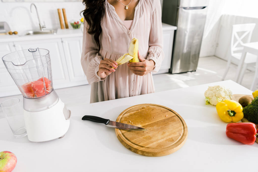 geschnittene Ansicht einer schwangeren Frau, die in der Nähe eines Mixers Banane mit Grapefruit schält  - Foto, Bild