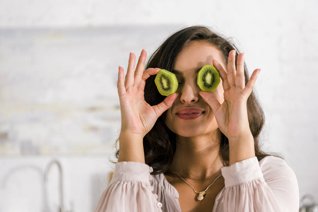 Szczęśliwa kobieta trzyma połówki owoców kiwi, jednocześnie pokrywając oczy  - Zdjęcie, obraz
