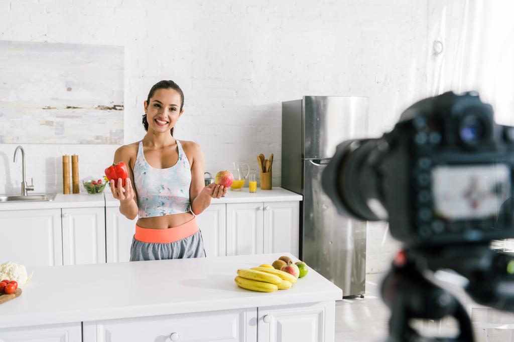 Selektiver Fokus eines glücklichen Mädchens mit Apfel und Paprika in der Nähe der Digitalkamera  - Foto, Bild